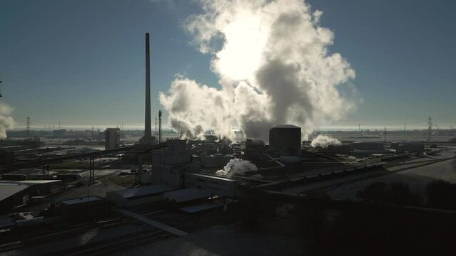 Smoke and steam rising of an industrial facility, CO2 emissions, air pollution, sunny, blue sky, aerial drone shot, backlit scene, slow movement up
