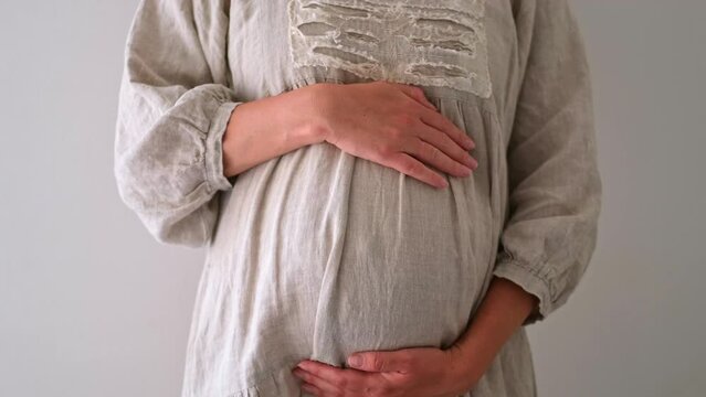 Unrecognizable pregnant woman caressing belly wearing linen beautiful dress,clothes on light grey background.Happy, healthy, stylish,fashion pregnancy.