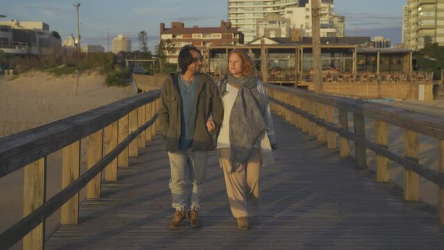 Pregnant couple holding hands walking by pier at sunset. Punta del Este Beach in Uruguay. 4K footage