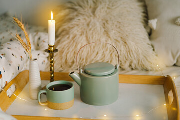 Obraz na płótnie Canvas Cup of tea and teapot on a serving tray on a bed. Breakfast in the bed in the morning. The concept of enjoying tea at home, spending time at home. A cozy autumn or winter concept.