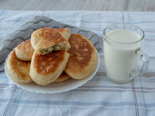 On table, covered with gray tablecloth, there is glass of fresh milk and plate with ruddy homemade pies, one pie is broken in half and one can see the filling of cottage cheese with herbs. Close-up