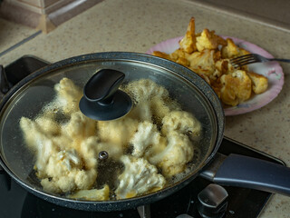 Pieces of cauliflower are fried in vegetable oil in a frying pan and there is a plate with fried golden cauliflower slices on the table. Concept of healthy and wholesome homemade food from vegetables