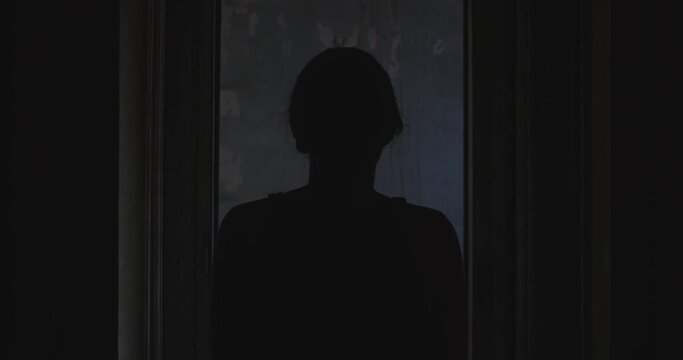 Black dark scene of a girl that walk away from behind and at the end she tilts her head making the atmosphere disquieting. The place is dark and spectral. Slow shot