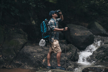 Hikers standing on the rock and use binocular to see animals and view landscape  with backpacks and background waterfall in the forest. hiking and adventure concept.