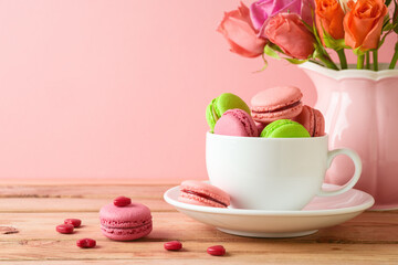 Fototapeta na wymiar Macaroons french cookies in coffee cup and rose flower bouquet over pink background. Happy Valentine's day concept.