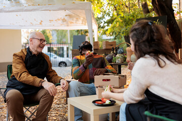 Group of cheerful people tasting food and wine at food fair, sitting together at table and having fun. Local farmers and clients enjoying glasses of homemade wine and fresh bio products.