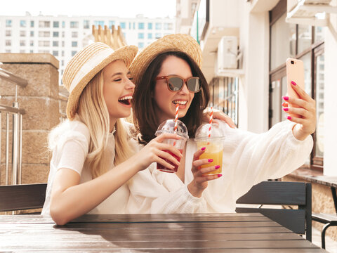 Two young beautiful smiling hipster female in trendy summer white dress and straw hats. Sexy carefree women holding and drinking fresh vegetable cocktail smoothie drink in plastic cup with straw