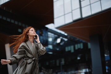 Woman walking around town and talking on the phone in trendy clothes against a backdrop of tall city buildings with an out-of-focus smile in windy weather