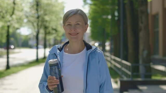 Portrait of happy mature female runner holding the sport bottle standing outside Senior woman in sportswear looking at the camera. Sport and healthy lifestyle concept.