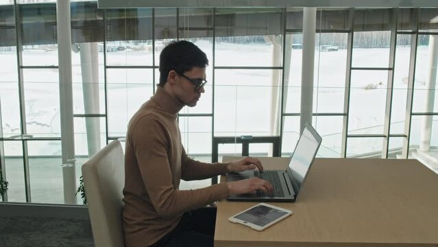 High angle side view of serious businessman in eyeglasses sitting at table working online using wireless devices