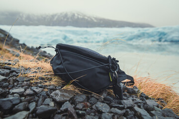 Close up camera bag on stones concept photo. Cameraman accessory outdoor. Front view photography with ice mountains on background. High quality picture for wallpaper, travel blog, magazine, article