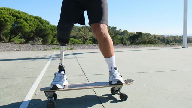 Closeup shot of man with leg prosthesis skateboarding outdoors. Athletic guy in shorts with disability doing sports on sunny day. Tracking shot. Amputee sport, motivation concept
