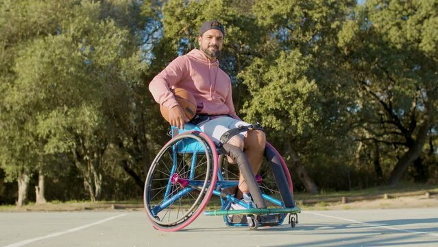 Happy Bearded Man In Sports Wheelchair At Basketball Court With Ball On His Lap, Taking It In Hand And Looking At Camera. Strong Man With Disability Doing Sports Outside. Amputee Sports Concept