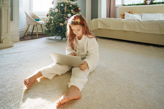 Girl wearing Christmas headband using laptop sitting on carpet at home