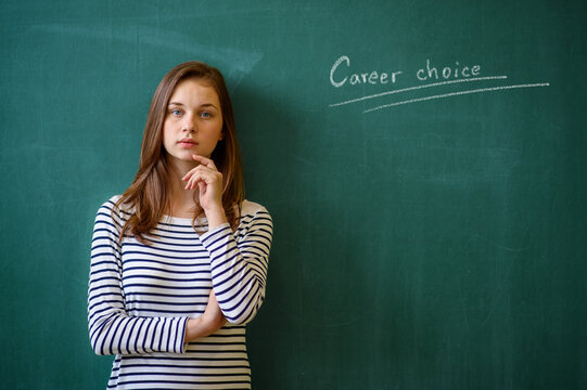 Career choice concept. Young female high school student standing in front of chalkboard in classroom, looking at camera.