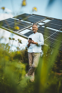 Mature man leaning on solar panels with smart phone at garden