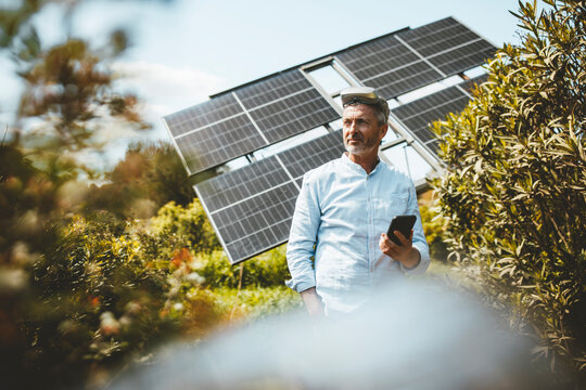 Smiling mature man standing with smart phone in front of solar panels