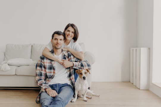 Portait of affectionate wife embraces husband who sits on floor near couch, their favourite pet poses at camera, make photo, being at home, buy new apartment, pose in living room with white walls