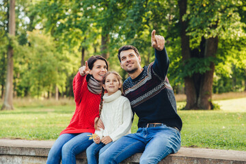 Fototapeta na wymiar Father and mother sit between their daughter against trees or nature background, show something to her, indicate with fore fingers. Smiling parents show bird to little beautiful girl. Parenthood