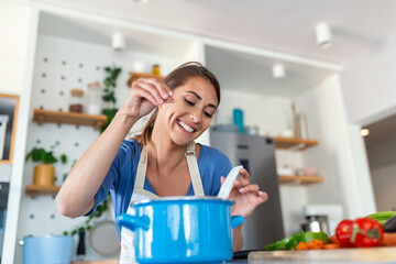 Happy Young Woman Cooking Tasting Dinner In A Pot Standing In Modern Kitchen At Home. Housewife Preparing Healthy Food Smiling . Household And Nutrition. Dieting Recipes Concept