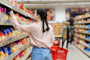 Back view of caucasian woman choosing products in grocery store. Shelves with food in background....