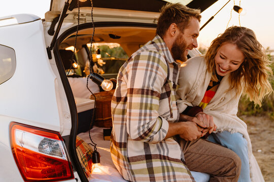 Happy Young White Couple Smiling And Sitting In Car Trunk Outdoors