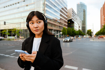 Fototapeta premium Young woman using cellphone and headphones while walking on street