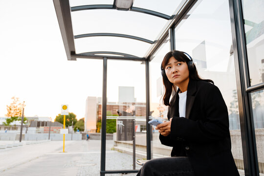 Young asian woman using cellphone and headphones sitting on bus station