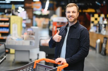 Customer In Supermarket. Man Doing Grocery Shopping Standing With Cart Choosing Food Product Indoors. Guy Buying Groceries In Food Store. Selective Focus, Copy Space.