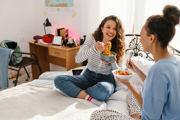 Two cheerful girls eating pasta while sitting on bed together