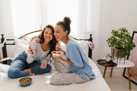 Two Cheerful Girls Eating Pasta And Using Mobile Phone While Sitting On Bed Together