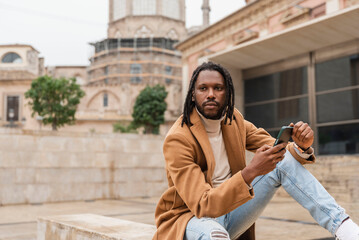 a stylish african american man with dreadlocks sitting in the city outdoors with smartphone in his hands