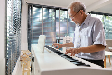 Happy smiling asian senior man with beard sitting and playing piano and singing a song in living...
