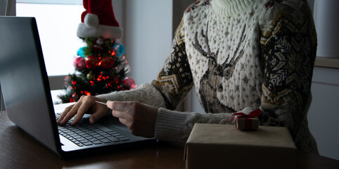 Woman holding credit bank card in hands involved in online shopping, purchasing Christmas gifts or ordering buying New year decorations in internet store using notebook. Santa`s hat. Christmas tree