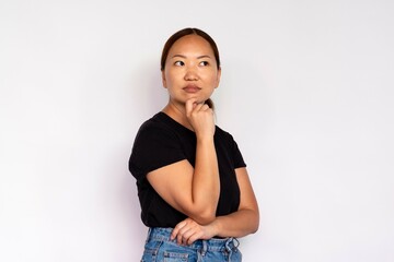 Portrait of concentrated young woman posing with hand on chin. Asian lady wearing black T-shirt and jeans standing and looking away over white background. Planning and thinking concept