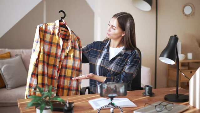 Smiling young female, recording video on camera her smart phone, fixed on tripod on desk. Showing clothes fashion. Say shopping opinion or promoting own brand store online. Marketing, advertising.