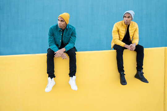 Funny black twin brothers wearing winter clothes, sitting on yellow and blue wall 