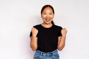 Portrait of excited young woman making winning gesture over white background. Asian lady wearing black T-shirt and jeans expressing success. Self confidence concept