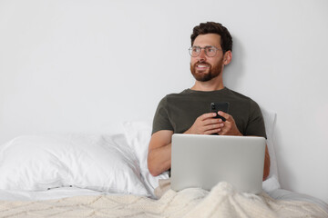 Handsome man using smartphone and laptop in bed, space for text