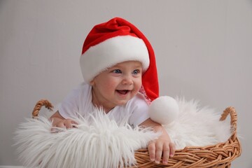 Cute baby wearing Santa hat in wicker basket on light grey background. Christmas celebration