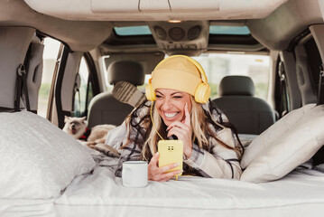 a laughing woman laying indoor mini camper van using a mobile phone and listening to the music with yellow headphones. Travel vanlife digital nomad people
