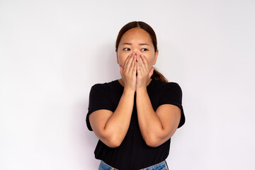 Portrait of scared young woman covering mouth with hands over white background. Asian lady wearing black T-shirt and jeans looking away frightened. Fear and stress concept