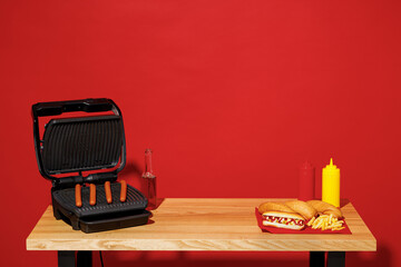 Wooden kitchen table with black electric grill sausage bottle beer kitchenware for making hotdog barbecue at cafe nobody isolated on plain red wall background studio. Cooking food bbq party concept.