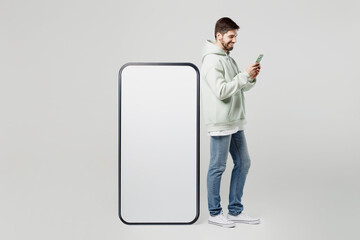 Full body happy young smiling fun man in mint hoody big huge blank screen mobile cell phone with workspace copy space mockup area hold use smartphone surfing isolated on plain solid white background