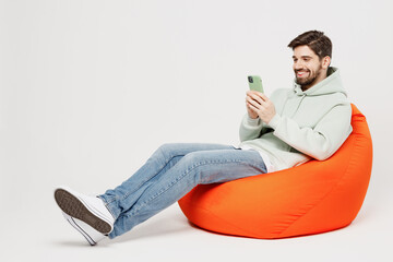 Full body young smiling happy fun cheerful man wear mint hoody sit in bag chair hold in hand use...