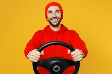 Young smiling happy fun cheerful cool caucasian man wear red hoody hat look camera hold steering wheel driving car isolated on plain yellow color background studio portrait. People lifestyle concept.