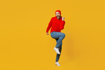 Fototapeta na wymiar Full body young happy fun caucasian man wear red hoody hat look camera doing winner gesture raise up leg clench fist isolated on plain yellow color background studio portrait People lifestyle concept