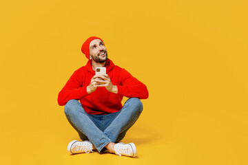 Full body minded young caucasian man wear red hoody hat look aside on workspace hold in hand use mobile cell phone isolated on plain yellow color background studio portrait. People lifestyle concept.