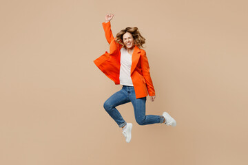 Full body fun young employee business woman corporate lawyer 30s wears classic formal orange suit glasses work in office jump high do winner gesture raise up hand isolated on plain beige background.