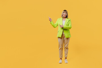 Full body elderly happy woman 50s years old wear green jacket white t-shirt point index finger aside indicate on workspace area copy space mock up isolated on plain yellow background studio portrait.